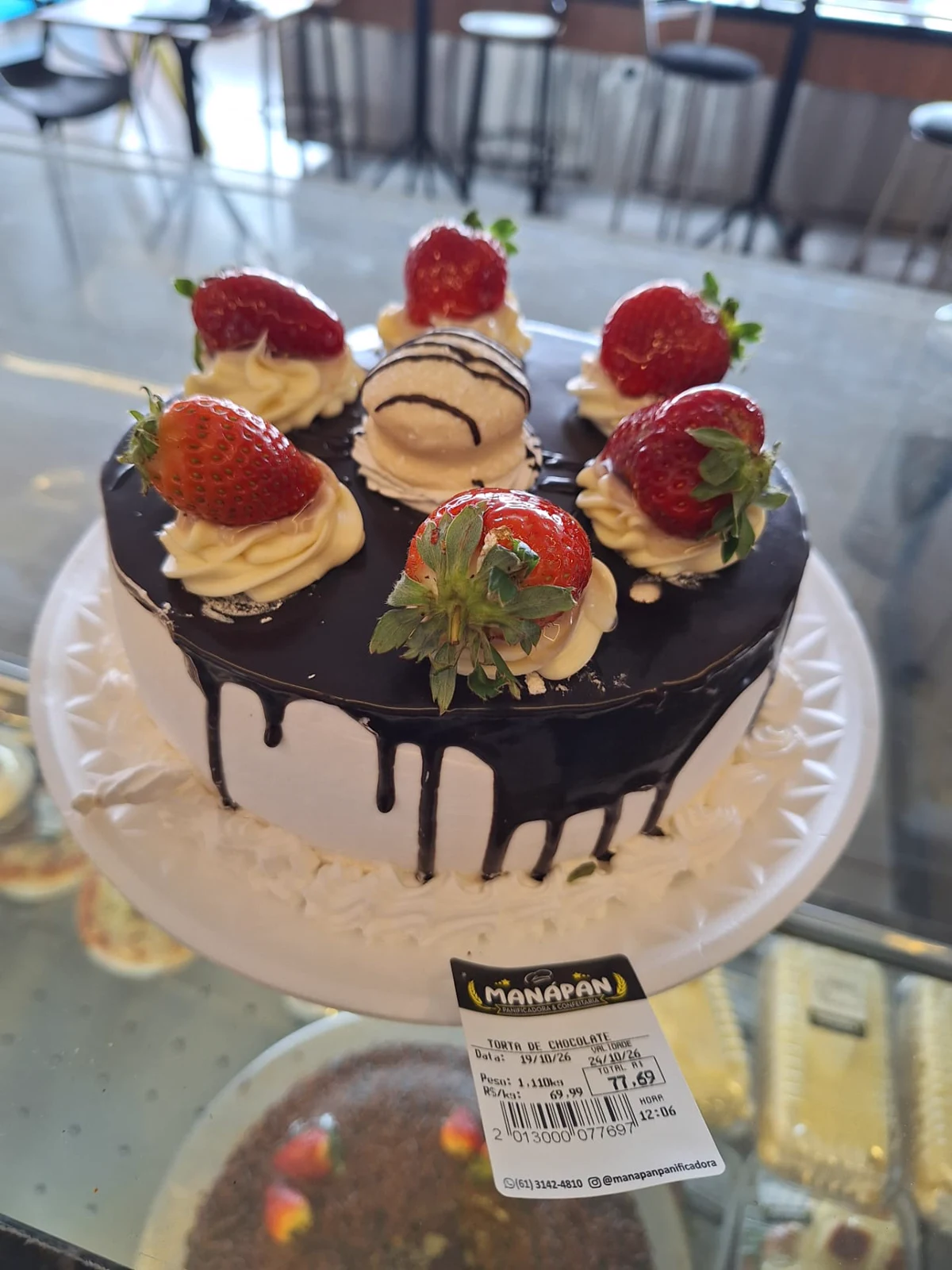 Torta de chocolate da ManáPan com ganache escorrido, chantilly e morangos, com etiqueta da padaria na vitrine