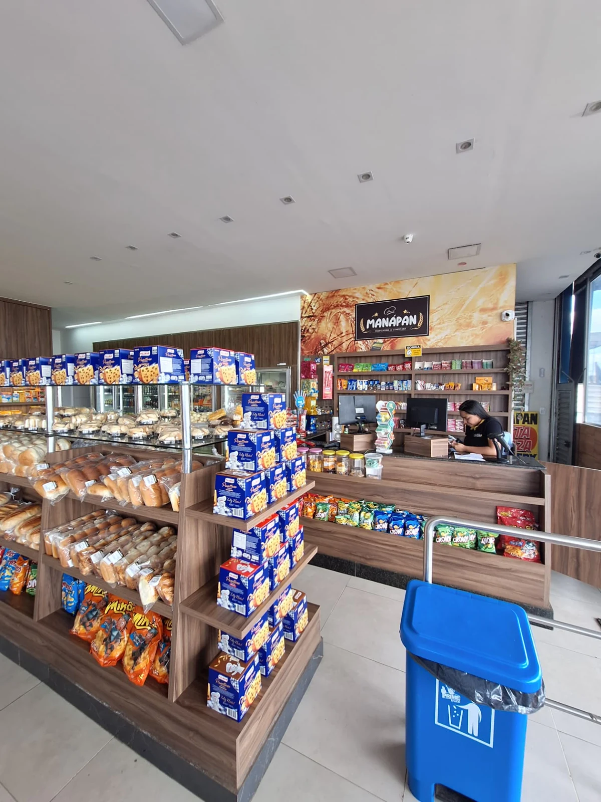 Área de caixa da ManáPan com atendente, prateleiras de pães embalados e produtos de conveniência