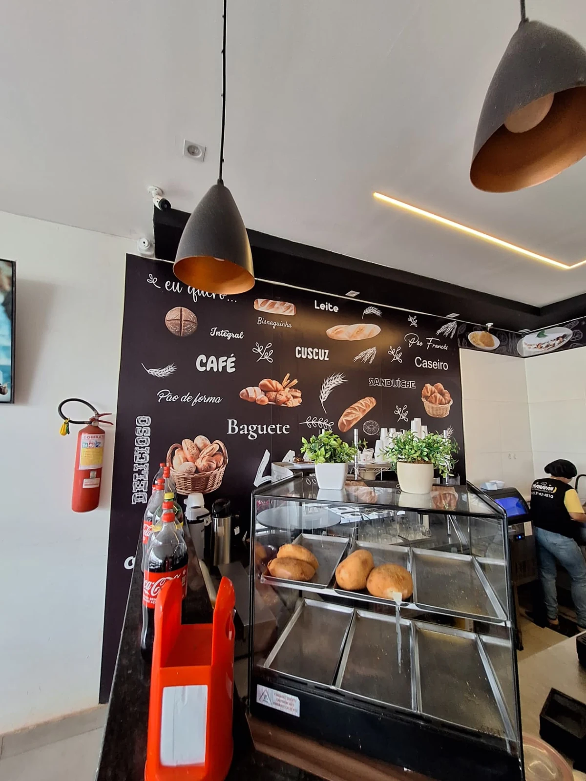 Balcão da ManáPan com vitrine de salgados, luminárias suspensas e parede decorada com pães